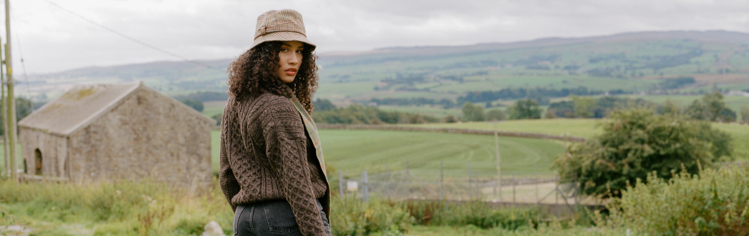 Woman standing in countryside in front of a barn and fields wearing a brown knitted aran jumper and tweed bucket hat