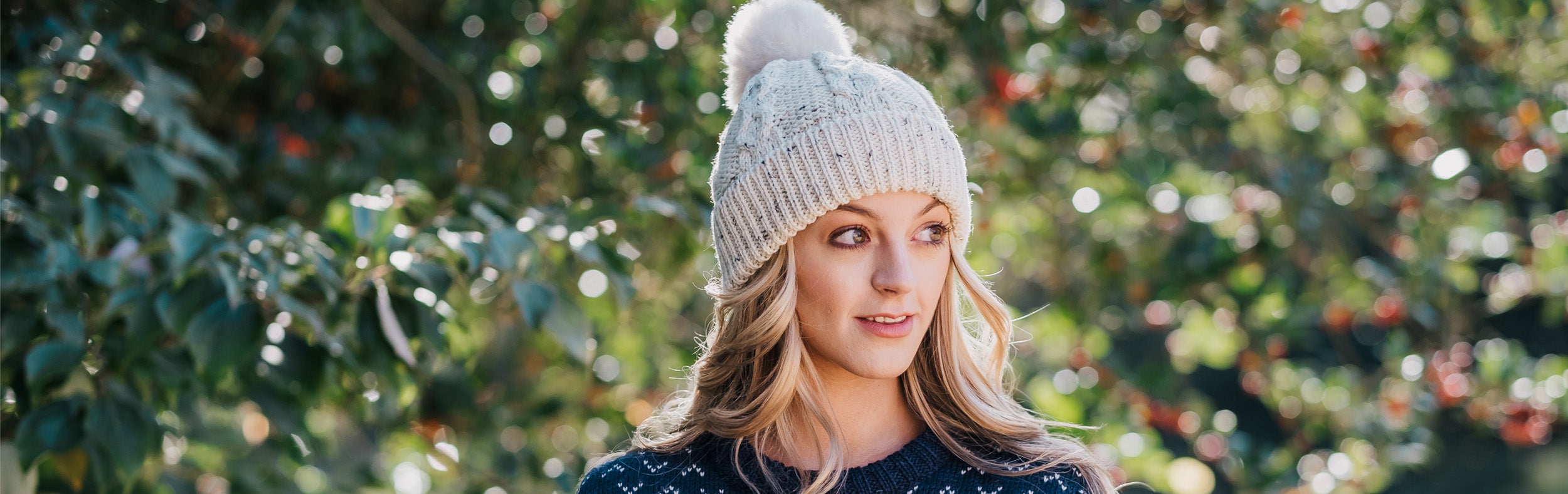 Woman wearing a knitted wool beanie with fluffy bobble, outside in front of green foliage