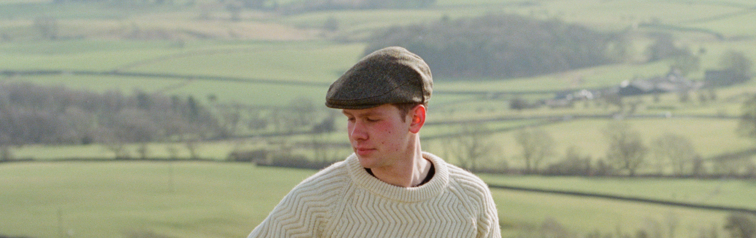 Photo of a man wearing a flat cap and knitted cream woolly jumper in front of a valley of fields and trees