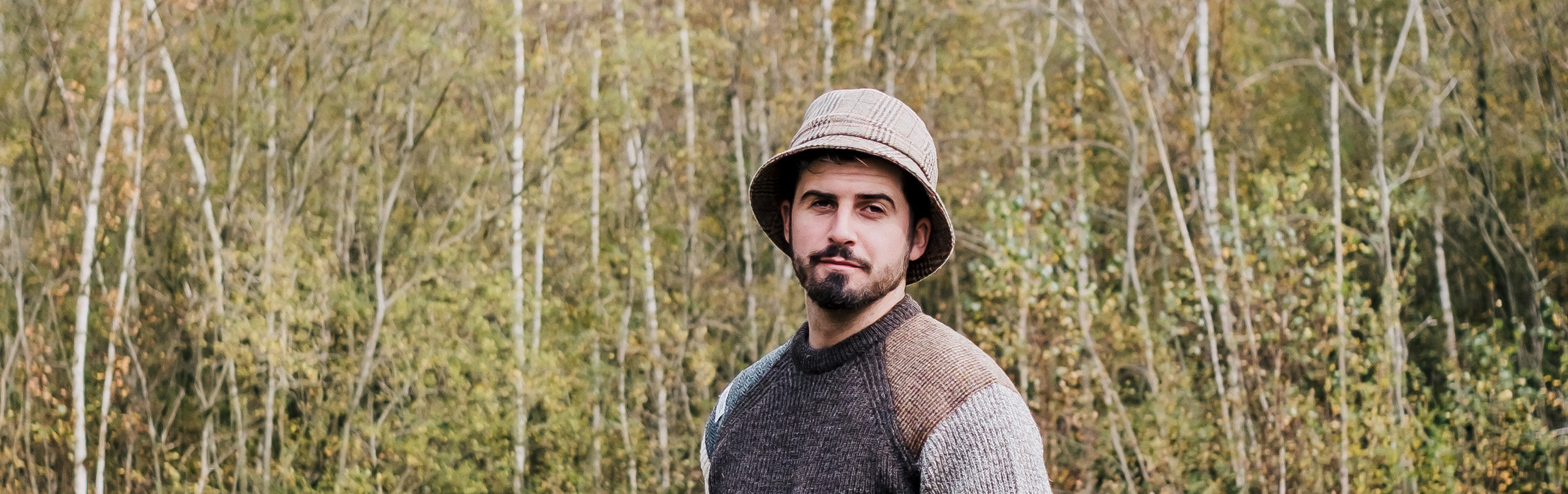 Man wearing a brown wool jumper with tweed shoulder patches and brown tweed bucket hat, standing in a forest