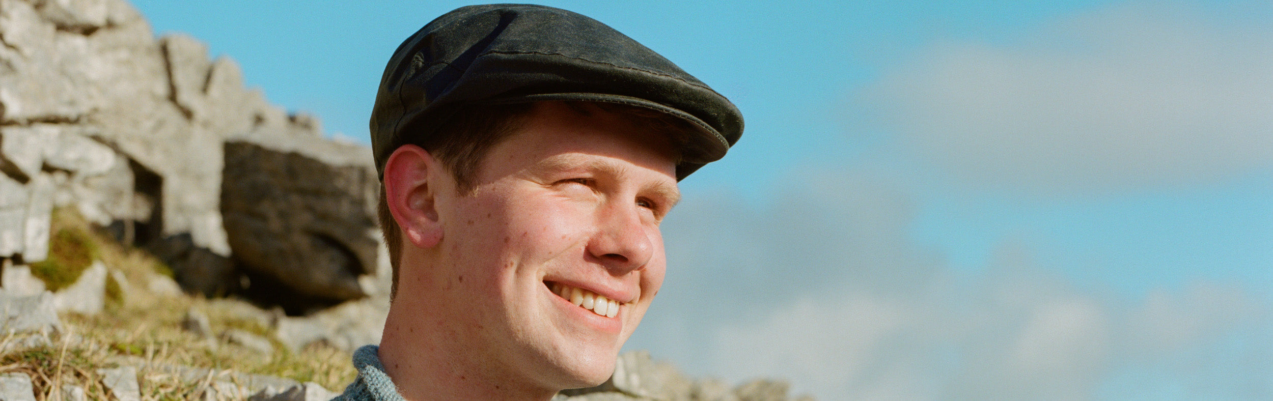 Close up of man wearing a wax flat cap with and outcrop of rocks behind him