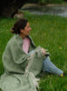 A woman wrapped in a wool blanket sitting on grass 