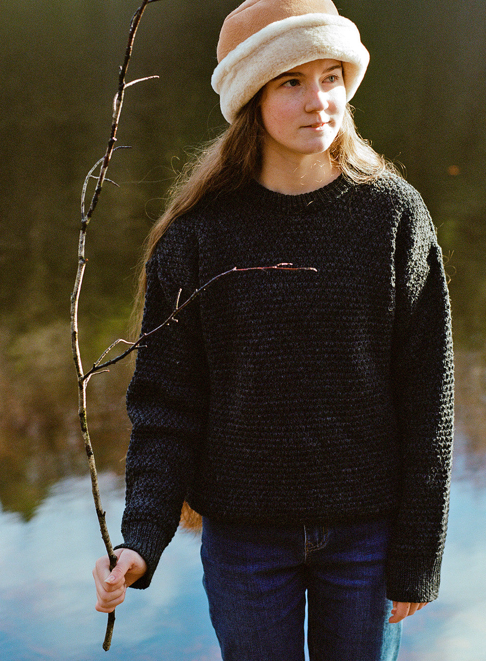 Girl wearing a tan colour sheepskin hat, charcoal knitted jumper and blue jeans, holding a stick in front of a lake