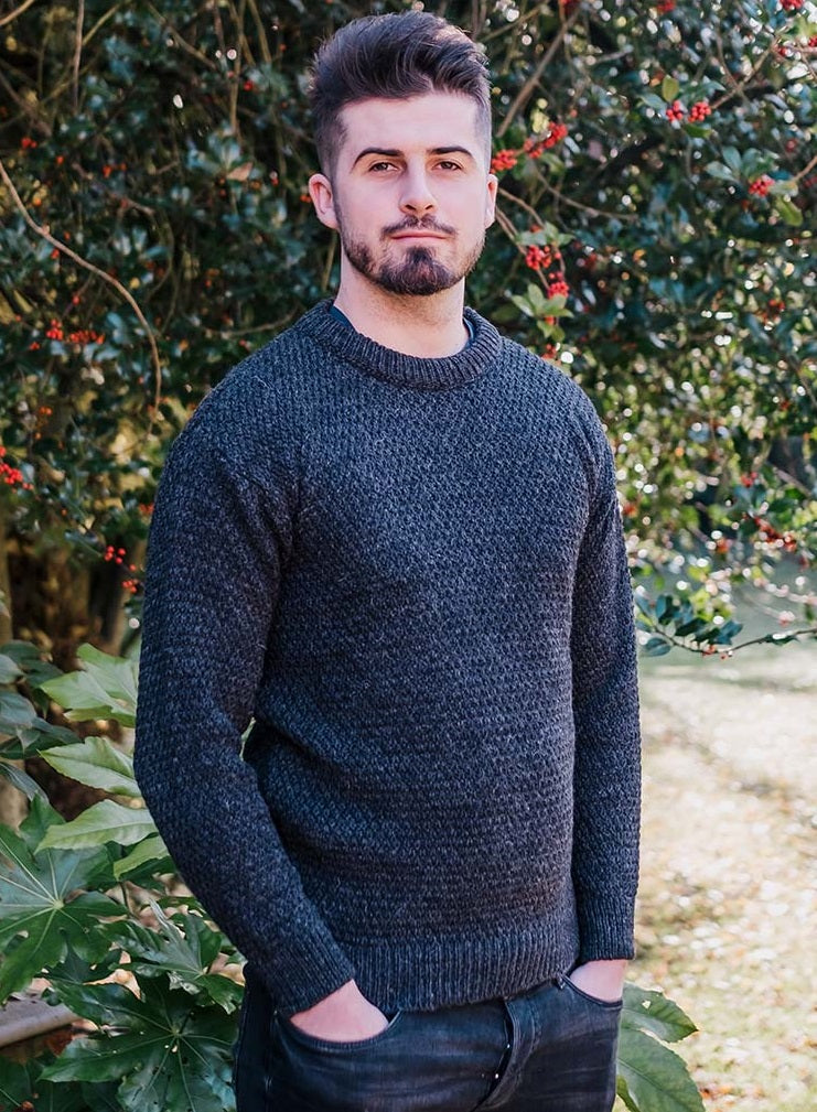 Man with beard wearing charcoal knitted jumper with hands in pockets of black jeans standing in front of a holly tree