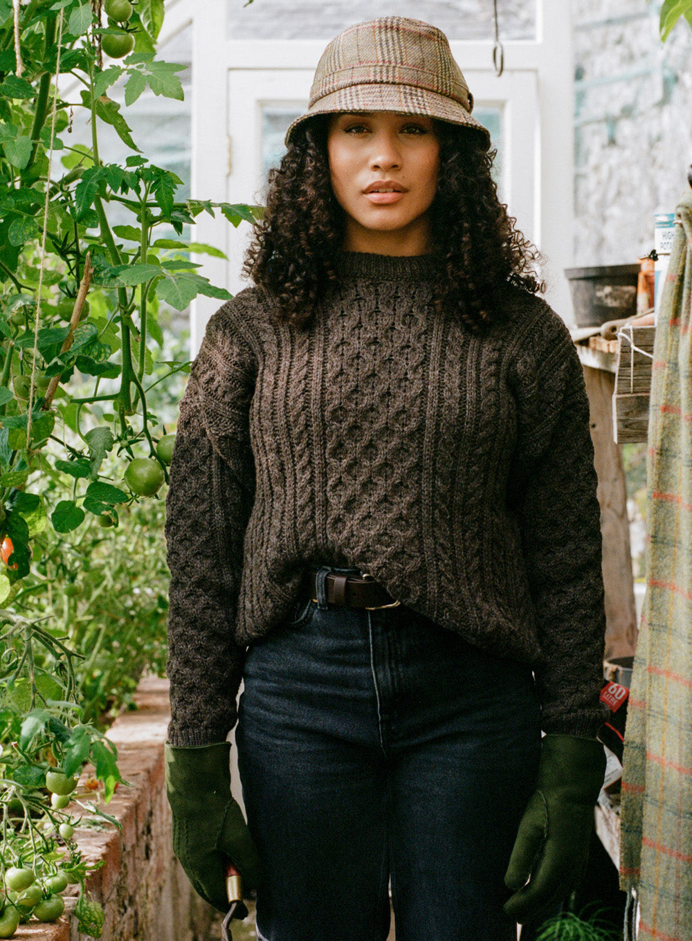 Female model wearing brown knitted aran jumper, brown tweed hat, jeans and sheepskin gloves next to a tomato vine in a greenhouse