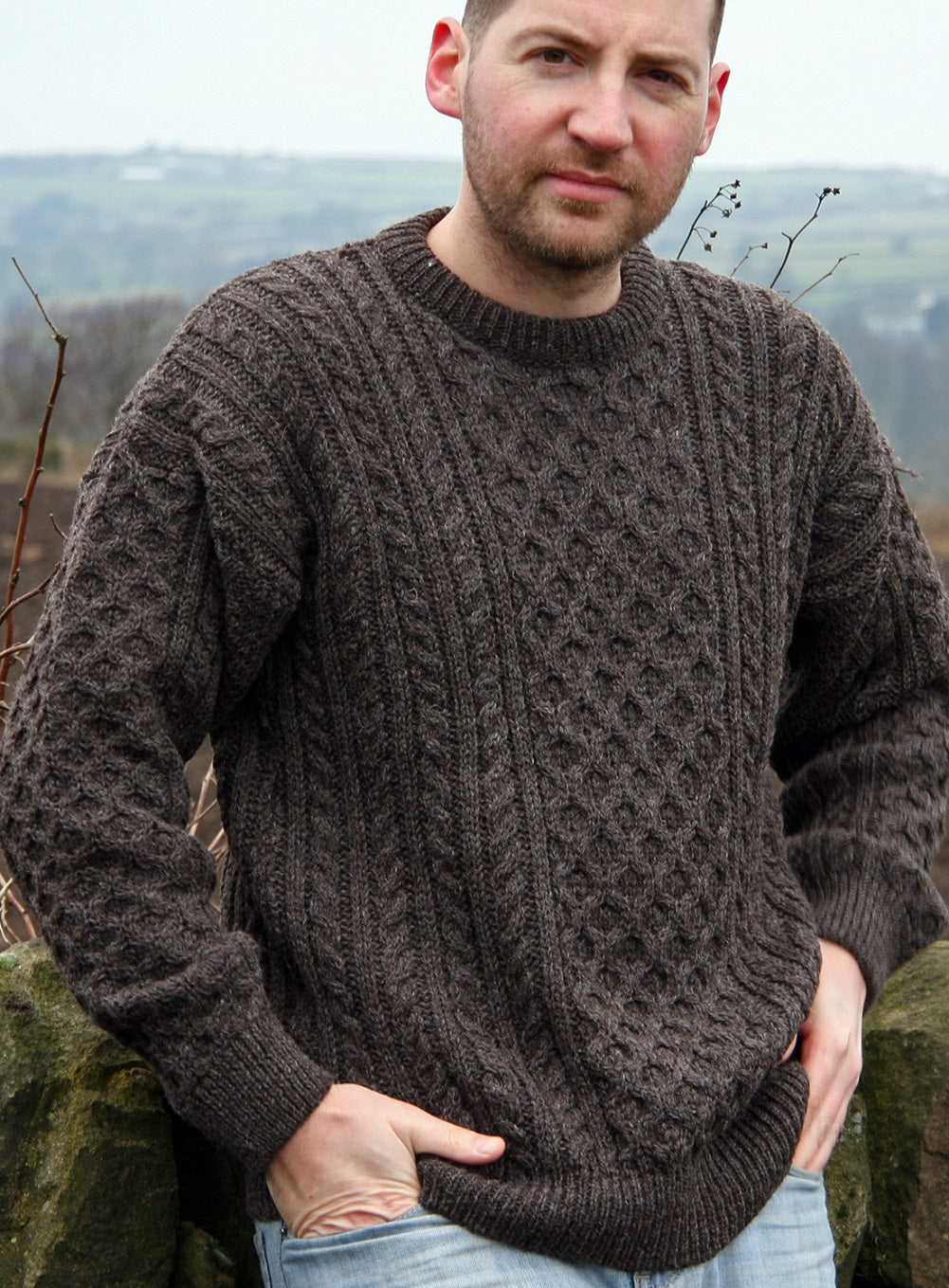 Male model wearing brown knitted Aran pattern jumper with hands in pockets of his jeans